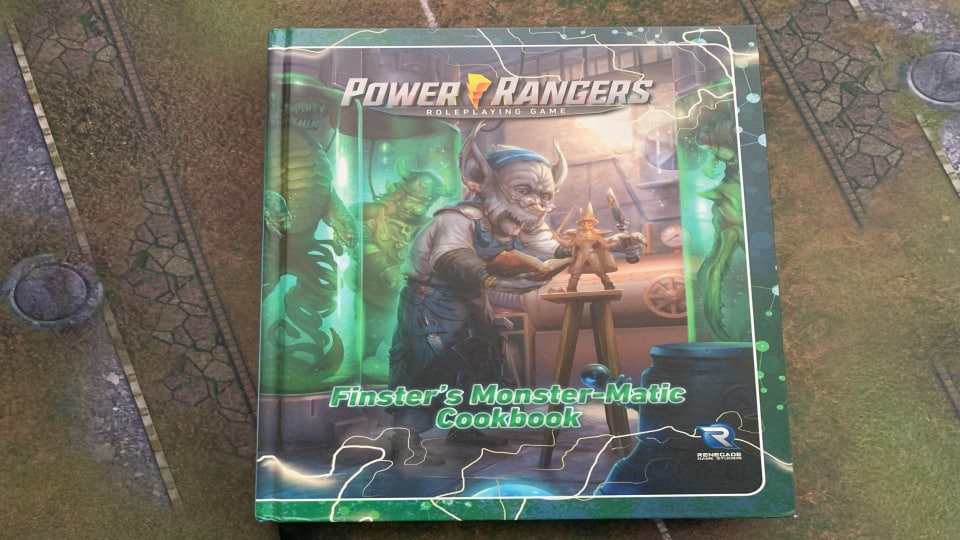 A hardcover copy of Finster's Monster-Matic Cookbook sitting on a table covered by a gaming mat.